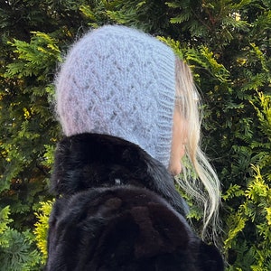 May include: A gray knitted hat with a diamond pattern. The hat is worn by a person with long blonde hair. The hat is pulled down over the person's ears.