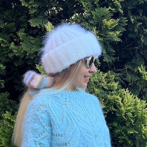 May include: A white fuzzy knit hat with a light blue sweater and a colorful knit hair tie.