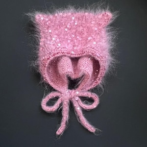 Puede incluir: Un gorro rosa y esponjoso hecho de hilo suave y texturizado, con pequeñas lentejuelas reflectantes dispersas por la superficie. El gorro presenta dos formas puntiagudas similares a orejas en la parte superior y largos lazos tejidos anudados en un lazo.