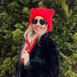May include: A bright red fuzzy knit hat featuring two pointed cat-like ears at the top and long dangling side ties. The hat has a soft, textured appearance and is worn with round black sunglasses that have thin gold-toned metal frames.