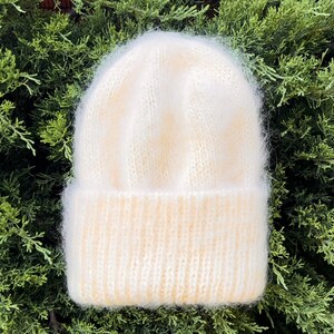 May include: A white fuzzy knit beanie hat with a ribbed brim. The hat is photographed against a green leafy background.