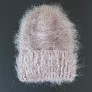 Op de afbeelding: Een licht mauve muts met een pluizige, langharige textuur. De muts heeft een geribbelde gebreide boord aan de basis en een gladde, ronde bovenkant. Het uiterlijk is zacht en volumineus door de lange, fijne vezels in het breiwerk.