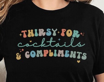 May include: A black t-shirt with the text "Thirsty for cocktails & compliments" in a rainbow of colors. The text is in a retro font style.