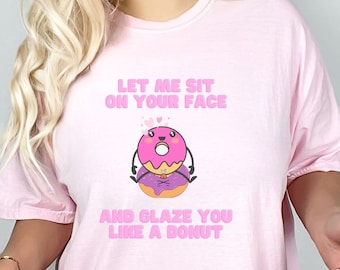 May include: A light pink t-shirt with the text "LET ME SIT ON YOUR FACE AND GLAZE YOU LIKE A DONUT" and a cartoon donut graphic. The donut has pink frosting, a purple bottom, and is smiling. The shirt is a Blossom color.