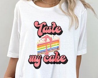 May include: White t-shirt with a colorful cake slice graphic and the text "Taste my cake" in pink and black. The cake slice features rainbow layers and pink frosting. A casual, short-sleeved tee.