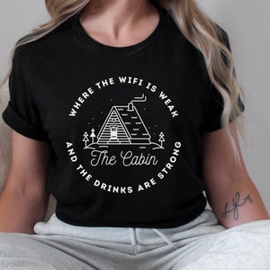以下が含まれることがあります： 白いキャビンと「Where the wifi is weak and the drinks are strong. The Cabin」というテキストが描かれた黒いTシャツ