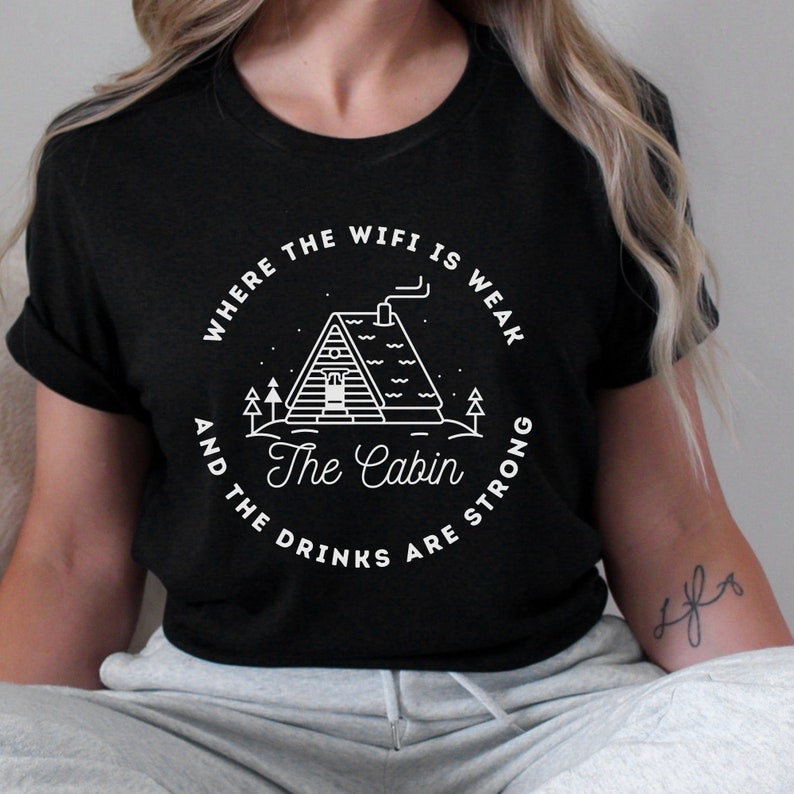 以下が含まれることがあります： 白いキャビンと「Where the wifi is weak and the drinks are strong. The Cabin」というテキストが描かれた黒いTシャツ