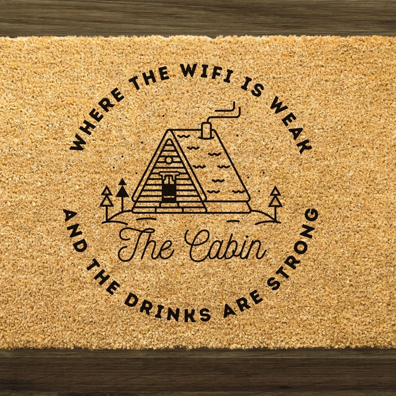 以下が含まれることがあります： 小屋のイラストの周りに円形で「Where the wifi is weak and the drinks are strong」という文字が書かれた茶色のドアマットです。