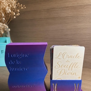 Peut inclure: Un jeu de cartes intitulé "L'Oracle du Souffle Divin" dans un support violet et bleu, à côté d'un support assorti avec le texte "l'origine de la lumiere" et "evelorys by vf". Les articles sont sur une surface en bois.