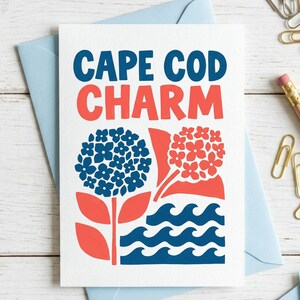 May include: A white greeting card with the text CAPE COD CHARM in blue and coral red. The design features a blue hydrangea, a coral red hydrangea, and stylized blue ocean waves at the bottom, all printed on a textured white cardstock surface.