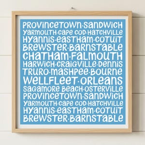 May include: A framed print with a light blue background and white text. The text lists various town names, including Provincetown, Yarmouth, and Chatham. The print is in a light brown wooden frame.
