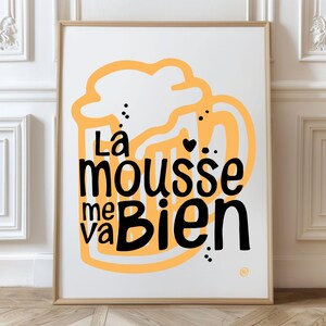 May include: Framed art print with a white background and a large orange beer mug illustration. The text "La mousse me va bien" is written in black, with a few black dots and a heart. The frame is a light brown color.