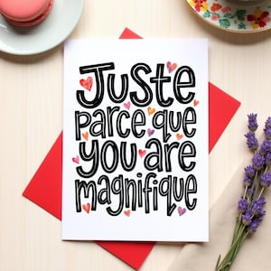 May include: A white greeting card with the text "Juste parce que you are magnifique" in black, decorated with small watercolor hearts. The card rests on a red envelope, with a sprig of lavender and a plate with a macaron in the background.