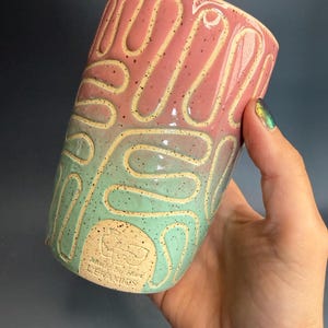 May include: A handmade ceramic tumbler with a two-tone design. The top half is pink, and the bottom half is mint green. A cream-colored, raised, maze-like pattern wraps around the exterior. The bottom of the tumbler has the text "Made by Hart Ceramics".
