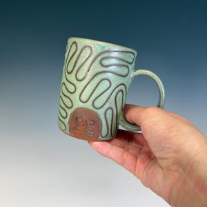 May include: A handmade ceramic mug with a light green glaze and brown squiggly lines. The mug has a rounded handle and a brown emblem near the base. The mug is held in a hand.