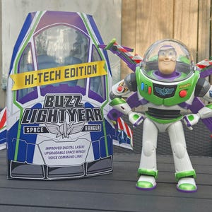 Op de afbeelding: Een Buzz Lightyear actiefiguur met een doorzichtige helm, paars en groen pak en uitgevouwen vleugels, naast de verpakking met de tekst HI-TECH EDITION, BUZZ LIGHTYEAR SPACE RANGER, IMPROVED DIGITAL LASER, UPGRADABLE SPACE WINGS, VOICE COMMAND LINK.