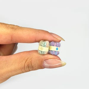 Puede incluir: Dos madejas de hilo en miniatura. Una es de color pastel con una banda amarilla y un punto beige. La otra es morada, azul y rosa con una banda amarilla y un punto azul. El hilo se sostiene entre los dedos.