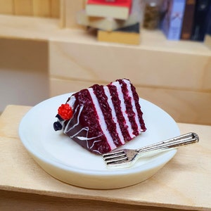 Può includere: Una fetta di torta Red Velvet in miniatura con glassa bianca e un lampone rosso in cima, servita su un piatto bianco con una forchetta argentata.
