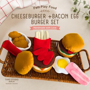 Op de afbeelding: Een vilten speelgoedvoedselset met een cheeseburger, een bacon-ei-burger, frietjes in een rode houder en kipnuggets in een witte kom. De set bevat een gebakken ei en een plakje spek, allemaal gerangschikt op een bruin gevlochten rieten dienblad.