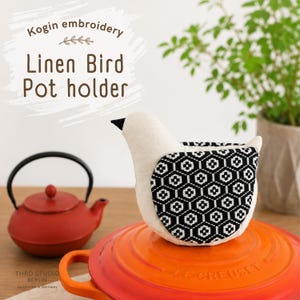 Könnte beinhalten: Ein vogelformiger Topflappen aus cremefarbenem Leinen mit einem schwarz-weißen geometrischen Kogin-Stickmuster auf dem Flügel. Der Vogel hat einen kleinen schwarzen Schnabel und steht auf dem Deckel eines leuchtend orangefarbenen Topfes.