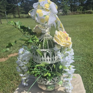 May include: A white metal birdcage centerpiece decorated with artificial yellow roses, white wisteria, and green ivy. The top of the cage is adorned with a large decorative ribbon featuring a yellow lemon pattern on a white background.