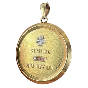 May include: A gold pendant with a textured finish. It features a cross design with small, clear stones and the words "QU'HIER QUE DEMAIN". The pendant has a loop at the top for a chain, and the date "4 AOU" is visible.