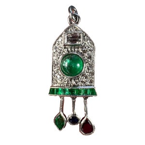 May include: A silver pendant with a large green emerald gemstone surrounded by smaller diamonds. The pendant has a rectangular shape with a pointed top and a green emerald trim. Three smaller gemstones hang from the bottom of the pendant: a green emerald, a blue sapphire, and a red ruby.
