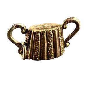 May include: A gold-colored, ornate miniature cup with two curved handles. The cup's body has a textured, carved design, and the handles are also intricately detailed. The cup is likely a decorative or collectible item.