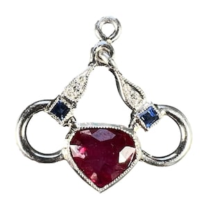 May include: A silver pendant featuring a heart-shaped ruby gemstone. The pendant has two blue square gemstones and small white gemstones. The design includes a loop at the top for a chain.