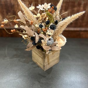 Könnte beinhalten: Eine Holzkiste gefüllt mit einem getrockneten Blumenarrangement. Die Anordnung enthält getrocknete Gräser, Blumen und Beeren in Braun-, Beige- und Schwarztönen.