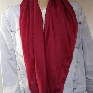 Pode incluir: Um cachecol vermelho escuro drapeado sobre os ombros de um manequim. O cachecol tem uma textura sedosa e é combinado com uma jaqueta jeans azul clara e uma camisa branca. A cor do cachecol é um tom rico e saturado.