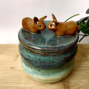 Op de afbeelding: Een ronde keramische pot met een deksel waarop twee kleine, bruine, gebeeldhouwde konijnenfiguren tegenover elkaar staan. De pot heeft een glanzend, meerkleurig glazuur dat verloopt van diep blauwgroen aan de bovenkant naar lichtgroen aan de basis.