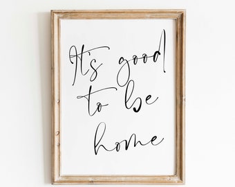 May include: A framed print with the text "It's good to be home" in black cursive script on a white background.