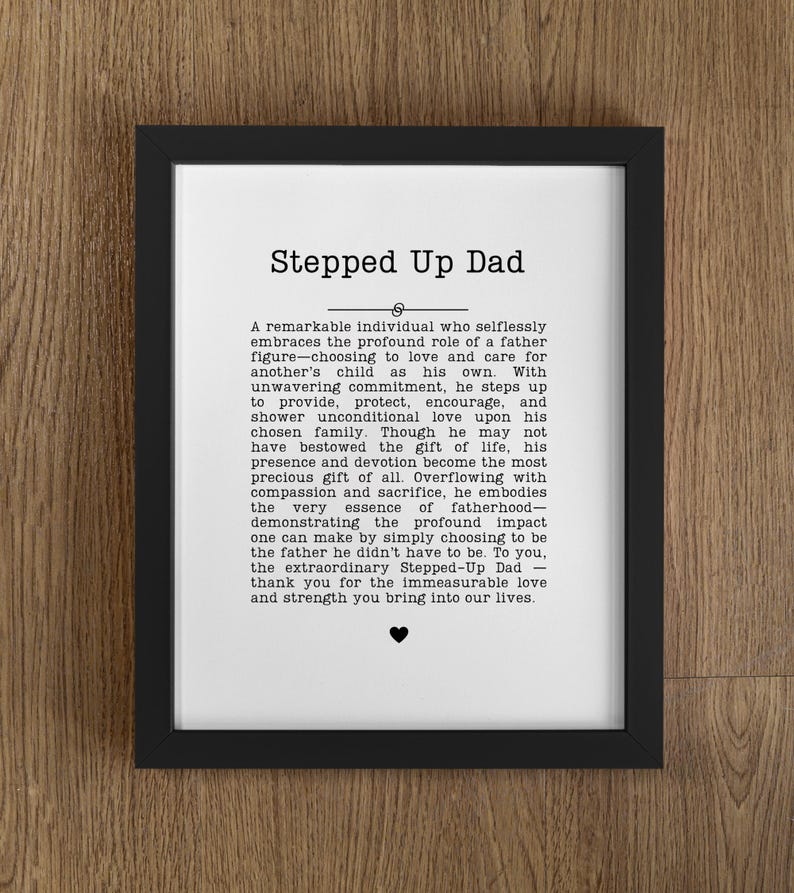May include: A framed print with the text "Stepped Up Dad" in a black frame. The print features a heartfelt message about a father figure, with a small heart symbol at the bottom. The background is a wooden surface.