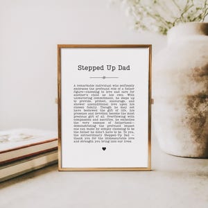 May include: A framed print with the text "Stepped Up Dad" in a gold frame. The text is a heartfelt message about fatherhood, with a small heart symbol at the bottom. The print is on a white background.