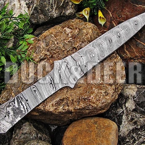 May include: A close-up of a silver knife blade with a Damascus steel pattern. The blade is sharp and pointed, and the pattern is intricate and detailed. The blade is lying on a bed of rocks.