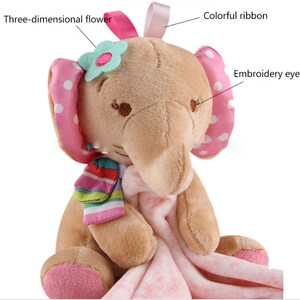 May include: A plush brown elephant toy with pink polka dot ears, a pink ribbon, and a three-dimensional flower. The elephant is holding a pink and white blanket.