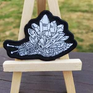 May include: A black and white embroidered patch of a snail carrying a pile of crystals on its back. The patch is displayed on a small wooden easel.