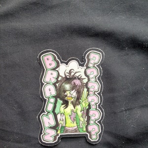 May include: A pink and green sticker with a cartoon zombie character with green skin, a peace sign, and the word "BRAINZ" in pink letters.