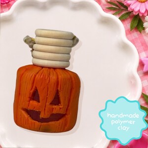 Può includere: Una figura di zucca in argilla polimerica arancione con volto intagliato e consistenza simile al legno, sormontata da un tappo cilindrico in legno chiaro a coste. Un'etichetta azzurra smerlata riporta il testo handmade polymer clay.
