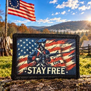 Könnte beinhalten: Ein rechteckiger Aufnäher mit schwarzem Rand, der einen Soldaten mit Dreispitz und Gewehr vor einem abgenutzten amerikanischen Flaggenmotiv zeigt. Der Text 1776 und STAY FREE ist in Weiß im unteren Bereich des Aufnähers aufgedruckt.