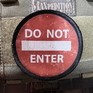 Puede incluir: Un parche circular rojo y negro con el texto "DO NOT ENTER" en letras blancas. El parche está unido a una bolsa de tela marrón y beige.