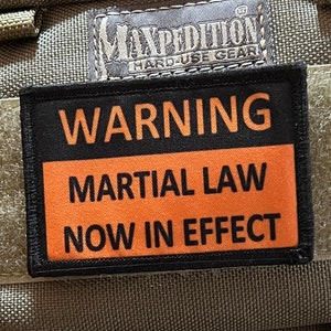 Op de afbeelding: Een groene tactische rugzak met een zwarte patch waarop "WARNING MARTIAL LAW NOW IN EFFECT" staat. De rugzak heeft een Maxpedition-logo op de voorkant.