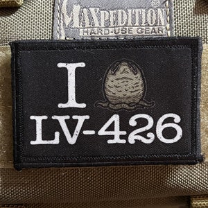 May include: A black patch with white text that reads "I LV-426" and a graphic of a creature from the movie Alien. The patch is attached to a tan Maxpedition bag.