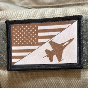 Może przedstawiać: Beżowo-szary plecak z czarną naszywką przedstawiającą flagę amerykańską i brązową sylwetkę myśliwca. Plecak ma czarną etykietę z napisem "Maxpedition Hard-Use Gear".