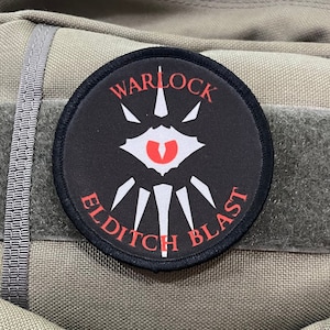 May include: A black and white patch with the text "Warlock Eldritch Blast" in white lettering on a black background. The patch has a red eye with white teeth and a white spike in the centre.
