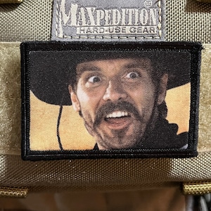 May include: A tan Maxpedition Hard-Use Gear backpack with a black patch featuring a man with a mustache and a surprised expression wearing a cowboy hat.