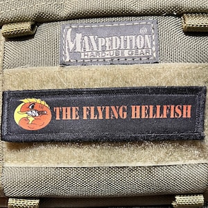 Peut inclure: Un sac à dos vert Maxpedition Hard-Use Gear avec un patch noir qui dit "THE FLYING HELLFISH" et un poisson de dessin animé avec un design rouge et orange.