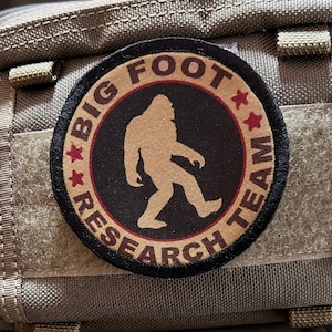 Puede incluir: Un parche negro y marrón con el texto "Big Foot Research Team" en un círculo con una silueta de Bigfoot caminando dentro del círculo. El parche está unido a una bolsa de tela beige.