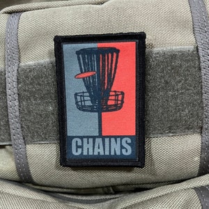 Può includere: Una toppa nera e grigia con un cestino da frisbee rosso e grigio e il testo "CHAINS" in bianco.